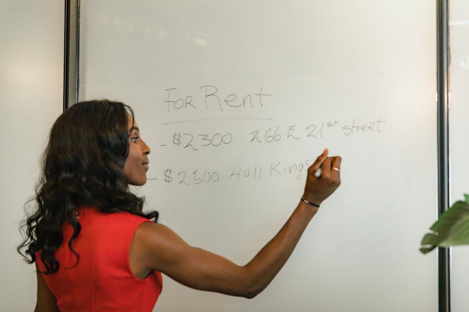 A woman with long, wavy black hair wearing a sleeveless red dress is writing on a whiteboard with a black marker. The whiteboard displays handwritten information about rental prices and addresses, inc