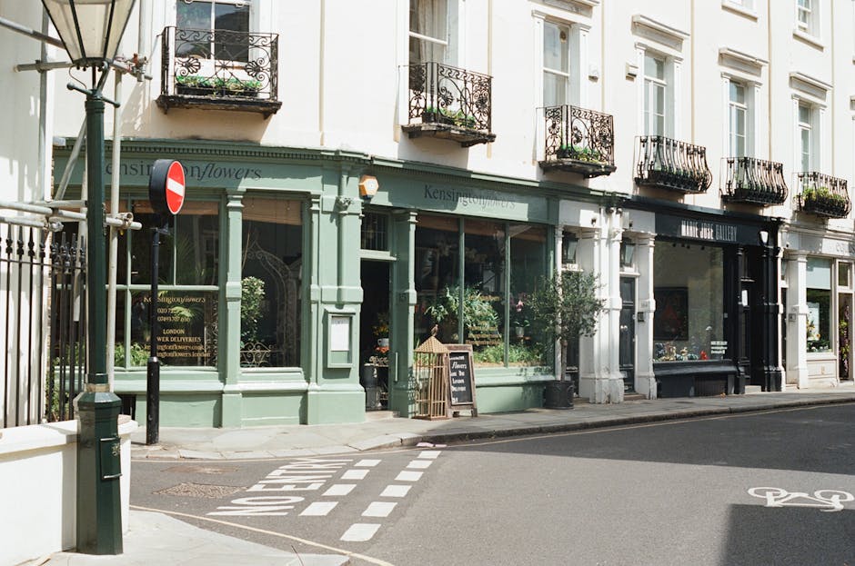 Exterior view of a flower shop located on a street corner with large display windows filled with fresh flowers. The shop is painted in a soft green color with white accents, featuring decorative trim 