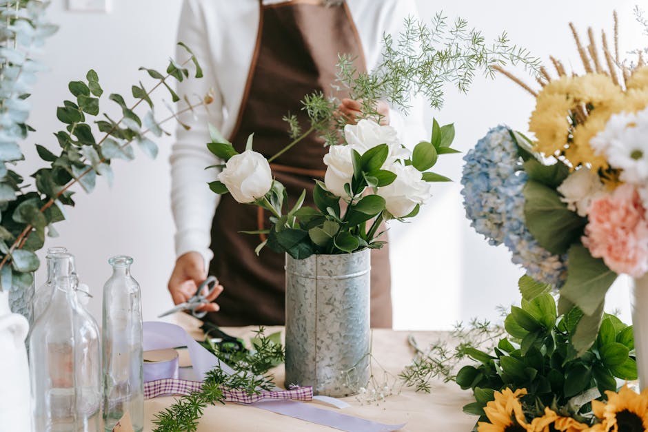 A person in a brown apron arranging a bouquet of white roses, green leaves, and delicate sprigs of foliage in a tall, textured silver vase on a light-colored table. Surrounded by various floral arrang