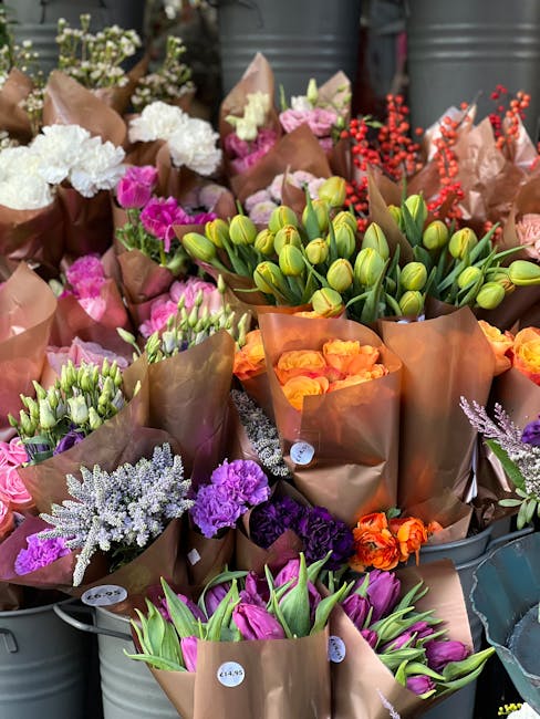 A variety of fresh flowers arranged in brown kraft paper and wrapped in cellophane, displayed outdoors. The bouquet includes white and pink carnations, purple lisianthus, orange roses, and green tulip