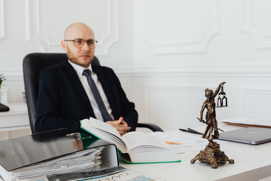 A professional florist in a formal office setting, sitting behind a desk with a large open book, surrounded by stacks of documents and a silver laptop. On the desk, there is a decorative bronze statue