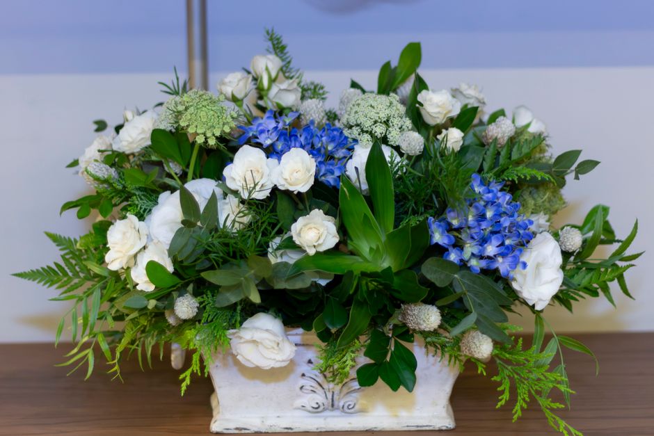 A floral arrangement featuring a mix of white roses and hydrangeas, accented with green foliage and ferns, arranged in a rectangular white ceramic vase. The bouquet is placed on a wooden surface again