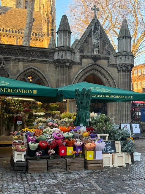 An outdoor flower stall situated on a cobblestone street in front of a historic stone building with gothic architectural features and trees with bare branches. The stall displays an array of fresh flo