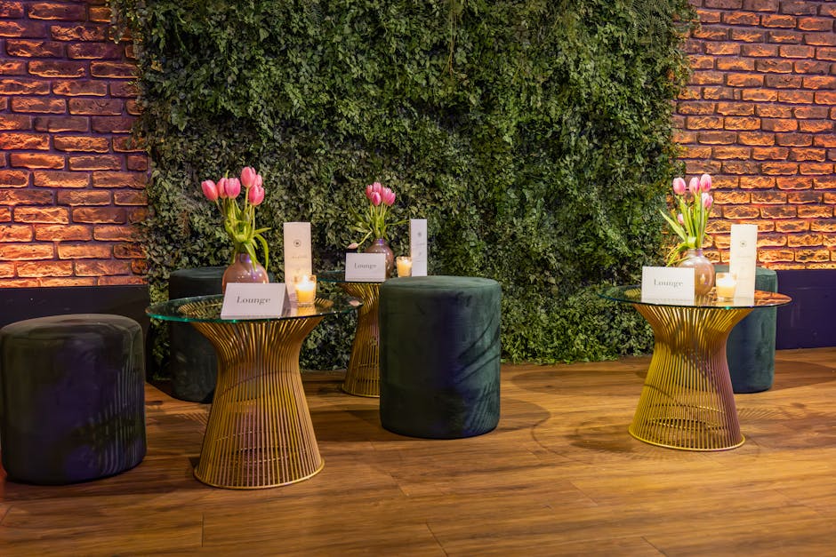 A stylish floral display featuring three small glass vases with pink tulips placed on round, gold-toned wireframe side tables in a lobby or lounge area. Each vase is paired with a white card labeled '