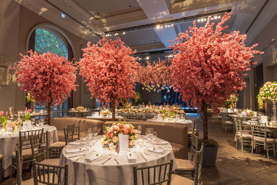 Elegant banquet hall decorated with three large cherry blossom trees with dense pink flowers, placed in grey pots, creating a striking centerpiece for a floral arrangement. The tables are covered with
