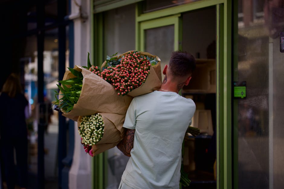 A person with short dark hair, wearing a light-colored T-shirt, is seen from behind holding a large, vibrant floral bouquet wrapped in brown craft paper. The bouquet contains clusters of pink and whit