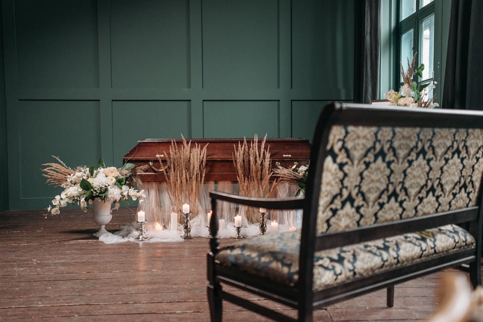 A wooden casket placed on the floor in a softly lit room with dark green paneled walls and large windows draped with black curtains. Surrounding the casket are floral arrangements, including white flo