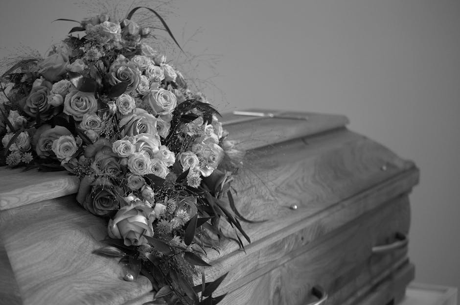 A large, fresh floral arrangement of roses and other flowers, predominantly in shades of light and dark roses, resting on the lid of a wooden casket with a natural wood grain finish. The bouquet inclu