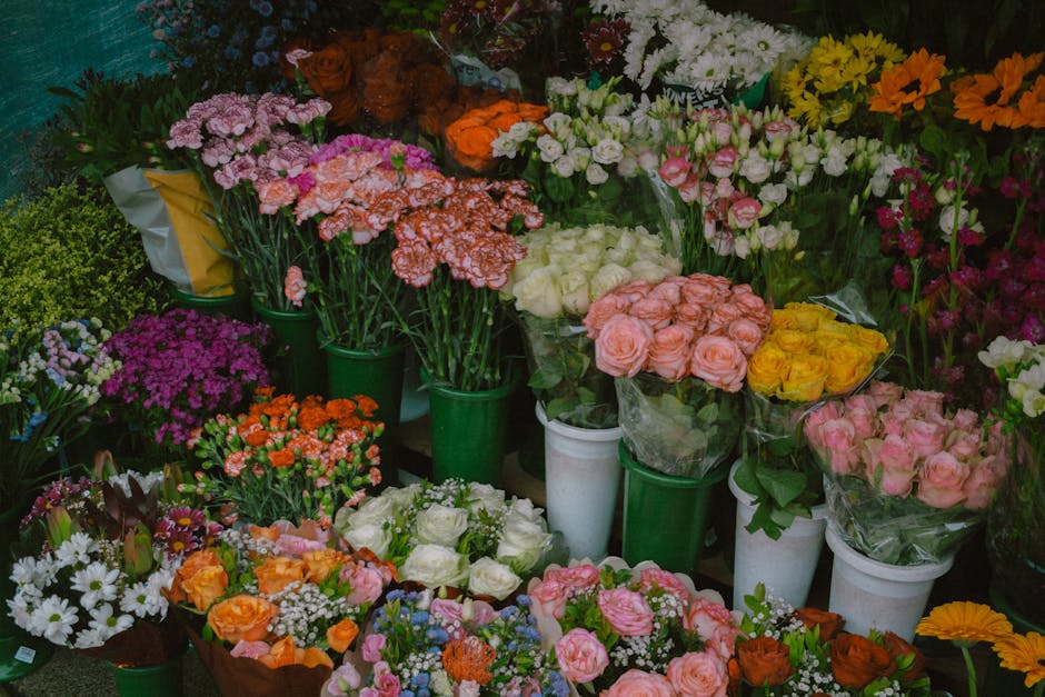 A diverse display of vibrant floral arrangements and bouquets placed in white and green vases, featuring roses in shades of pink, peach, yellow, and white, as well as carnations, daisies, chrysanthemu